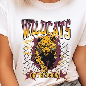 May include: White t-shirt featuring a graphic of a snarling wildcat bursting through a chain-link fence. The word "WILDCATS" is arched above the image in yellow and maroon, with "ON THE PROWL" below in a starburst banner.