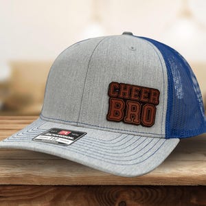 May include: A gray trucker hat with a blue mesh back and a curved brim. The hat features the text "CHEER BRO" in brown lettering on a brown patch. The hat is sitting on a wooden surface.