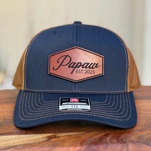 May include: A navy blue and brown trucker hat with a leather patch. The patch has the word "Papaw" in script and "EST 2025". The hat has a curved brim and a mesh back. The brand tag reads "Richardson 112".