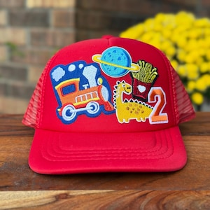 May include: Red trucker hat with a mesh back and a curved brim. The front features colorful embroidered patches of a train, a planet, fries, a dinosaur, and the number 2. The hat is likely for a child.