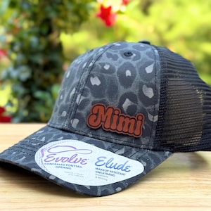 May include: A gray and black trucker hat with a leopard print pattern. The hat features a brown leather patch with the name "Mimi" in orange lettering. The hat has a black mesh back and a curved brim.