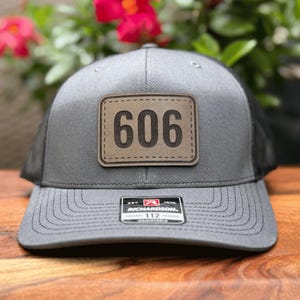 May include: Gray trucker hat with a black mesh back and a curved brim. A rectangular patch with the number "606" is centered on the front. The hat has a Richardson 112 tag.