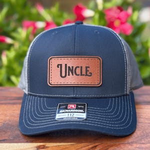 May include: Navy blue trucker hat with a leather patch that reads "UNCLE" in a serif font. The hat has a gray mesh back and a curved brim. The Richardson 112 label is visible on the front.