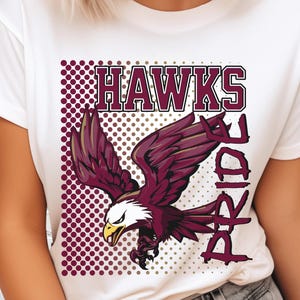 May include: White t-shirt featuring a maroon eagle graphic with the word "HAWKS" above and "Pride" to the side. The eagle has a white head, yellow beak, and maroon wings, set against a dotted background.