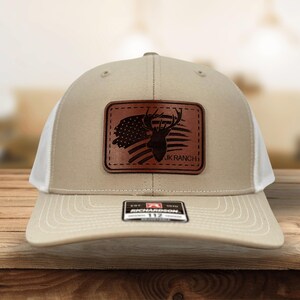 May include: A beige and white trucker hat featuring a rectangular brown leather patch on the front. The patch displays a silhouette of a deer head overlaid on an American flag design with the text JK RANCH. A black sticker is on the brim.