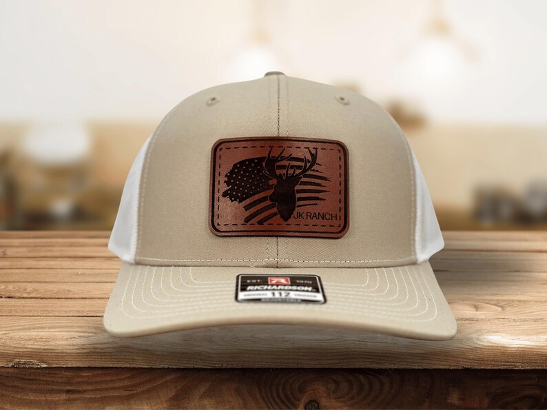 May include: A beige and white trucker hat featuring a rectangular brown leather patch on the front. The patch displays a silhouette of a deer head overlaid on an American flag design with the text JK RANCH. A black sticker is on the brim.