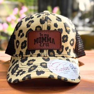 May include: A baseball cap with a tan and black leopard print pattern, featuring a brown rectangular leather patch on the front with the text In my MOMMA Era embossed in black. The cap has black mesh side panels and a circular white product tag.