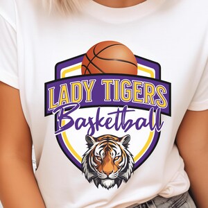 May include: White t-shirt featuring a basketball and a tiger head graphic. The design includes the words "LADY TIGERS Basketball" in purple and yellow. The basketball is brown and the tiger has orange and black stripes.