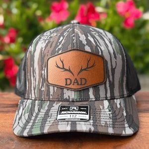 May include: Camouflage trucker hat with a brown leather patch featuring the word "DAD" and deer antlers. The hat has a black mesh back and a curved brim. The brand "Richardson 112" is visible on the front.
