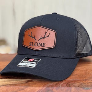 Pu&ograve; includere: Un cappellino da baseball nero con retro in rete e cinturino regolabile. La parte anteriore presenta una toppa in similpelle marrone con una grafica nera di corna di cervo e il testo SLONE. Sulla visiera &egrave; presente un adesivo bianco e nero con Richardson 112.