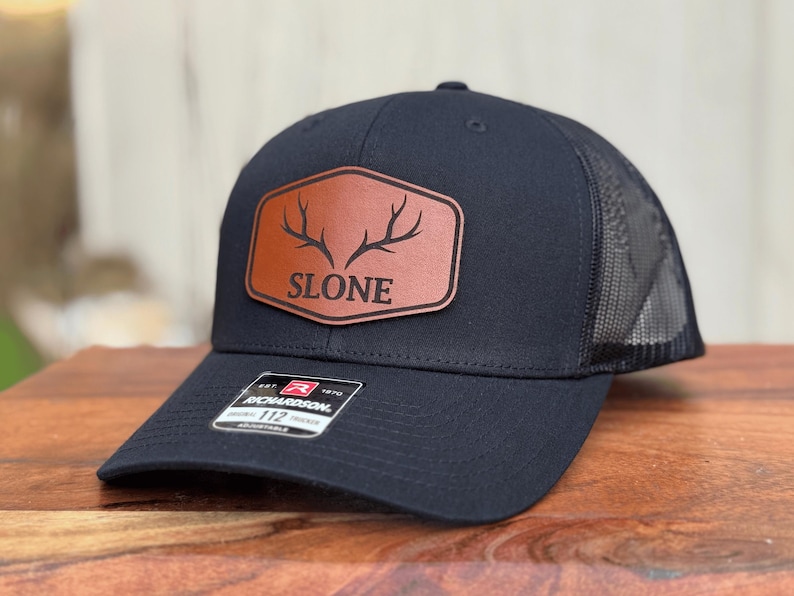 Pu&ograve; includere: Un cappellino da baseball nero con retro in rete e cinturino regolabile. La parte anteriore presenta una toppa in similpelle marrone con una grafica nera di corna di cervo e il testo SLONE. Sulla visiera &egrave; presente un adesivo bianco e nero con Richardson 112.