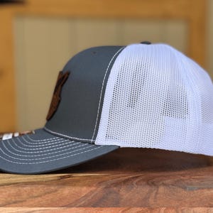 May include: A grey baseball cap with a white mesh back and a curved brim. The front panel features a brown leather-like patch. The cap has white stitching along the brim and crown, with a small rectangular tag attached to the side of the brim.