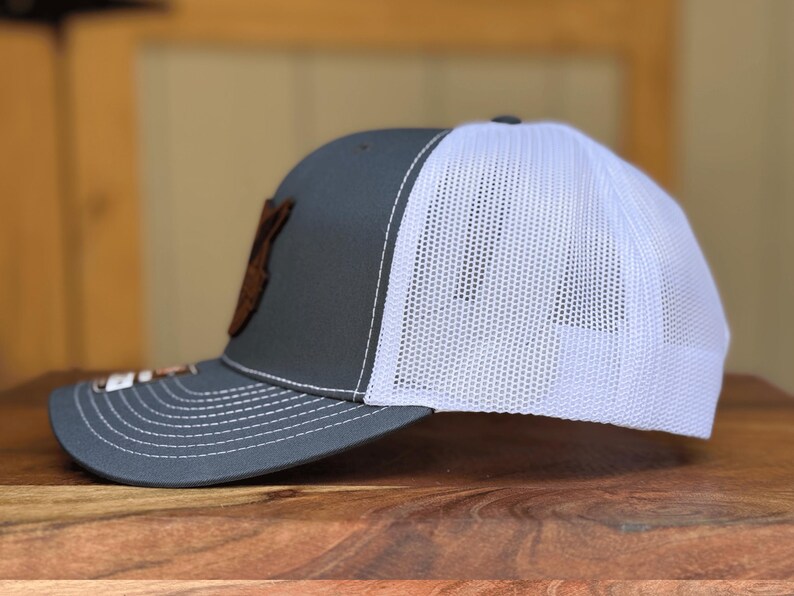 May include: A grey baseball cap with a white mesh back and a curved brim. The front panel features a brown leather-like patch. The cap has white stitching along the brim and crown, with a small rectangular tag attached to the side of the brim.