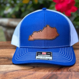 May include: A blue and white trucker hat with a brown leather patch of the state of Kentucky. The hat has a curved brim and an adjustable strap. The Richardson brand label is visible.
