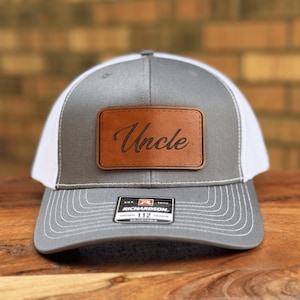 May include: A grey and white trucker hat featuring a rectangular brown leather patch on the front panel. The patch is embossed with the word Uncle in a black script font. A black and white sticker is centered on the brim with Richardson 112 text.