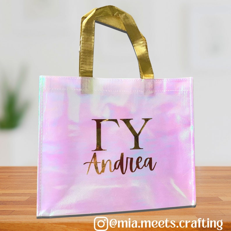 K&ouml;nnte beinhalten: Eine rosa schimmernde wiederverwendbare Einkaufstasche mit goldenen Henkeln und dem in Gold gedruckten Text "&Gamma;&Upsilon; Andrea".