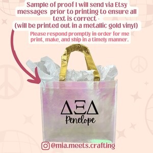 K&ouml;nnte beinhalten: Eine wei&szlig;e, schimmernde Tragetasche mit goldenen Henkeln. Die Tasche hat ein schwarzes Vinyl-Design mit den griechischen Buchstaben Delta, Epsilon, Delta und dem Namen Penelope. Die Tasche steht auf einem rosa und wei&szlig;en Hintergrund.