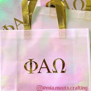 K&ouml;nnte beinhalten: Eine wei&szlig;e Tragetasche mit irisierendem Schimmer und goldenen Henkeln. Die Tasche hat die griechischen Buchstaben "&Phi;&Alpha;&Omega;" in Gold auf der Vorderseite aufgedruckt. Der Instagram-Handle "@mia.meets.crafting" ist unter den Buchstaben aufgedruckt.