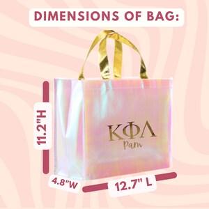 K&ouml;nnte beinhalten: Eine rosa irisierende Tragetasche mit goldenen Henkeln und den griechischen Buchstaben "&Kappa;&Phi;&Lambda;" sowie dem Namen "Pam" in Gold gedruckt. Die Tasche misst 11,2 Zoll in der H&ouml;he, 12,7 Zoll in der Breite und 4,8 Zoll in der Tiefe.