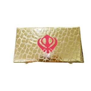 May include: A rectangular, gold-coloured box with a textured, crocodile-like pattern. A red Khanda symbol is centered on the top. The box has a shiny, metallic finish and appears to be made of a rigid material.