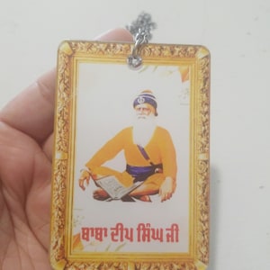 May include: A gold-framed picture of a man with a white beard and turban, holding a book. The picture is on a white background and has the text "Baba Sri Chand Ji" in red.
