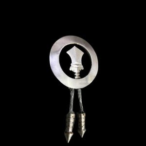 Puede incluir: Una corbata bolo plateada con un colgante circular. El colgante presenta un diseño central que se asemeja a una corona o punta de lanza. Dos puntas plateadas cuelgan del colgante, conectadas por cordones trenzados.