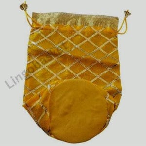 Puede incluir: Bolsa amarilla con cordón, diseño de rombos y detalles dorados. La bolsa tiene una base redonda y una parte superior dorada. El cordón es amarillo con extremos decorativos dorados. Para transportar objetos pequeños.
