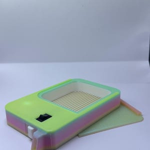 K&ouml;nnte beinhalten: Ein rechteckiges Ger&auml;t in Neongr&uuml;n und Pastellt&ouml;nen mit einer kleinen rechteckigen &Ouml;ffnung und einem Schalter. Das Innere ist hellgr&uuml;n mit parallelen Linien. Ein kleines, flaches, rechteckiges St&uuml;ck liegt neben dem Ger&auml;t.