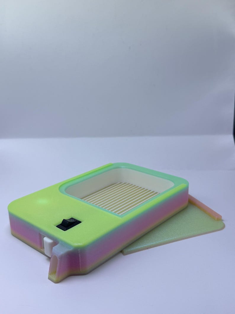 K&ouml;nnte beinhalten: Ein rechteckiges Ger&auml;t in Neongr&uuml;n und Pastellt&ouml;nen mit einer kleinen rechteckigen &Ouml;ffnung und einem Schalter. Das Innere ist hellgr&uuml;n mit parallelen Linien. Ein kleines, flaches, rechteckiges St&uuml;ck liegt neben dem Ger&auml;t.