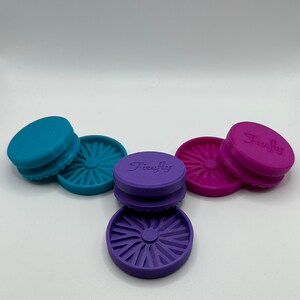 Puede incluir: Tres tapas redondas de silicona con un dise&ntilde;o de estrella. Las tapas son de color turquesa, morado y rosa. La tapa morada tiene la palabra "Firefly" impresa.