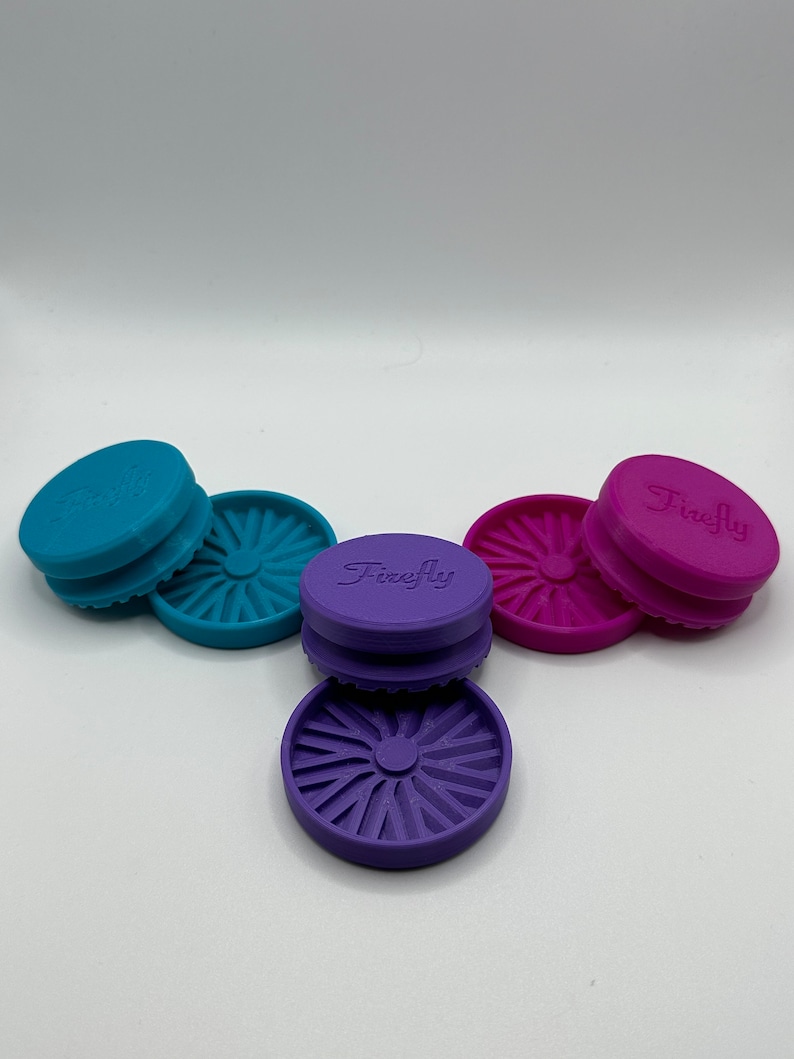 Puede incluir: Tres tapas redondas de silicona con un dise&ntilde;o de estrella. Las tapas son de color turquesa, morado y rosa. La tapa morada tiene la palabra "Firefly" impresa.