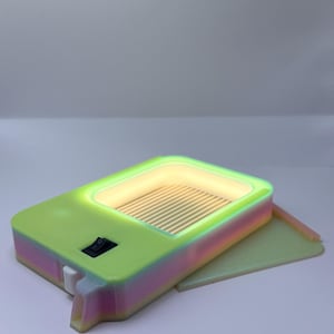 K&ouml;nnte beinhalten: Ein neongr&uuml;nes und regenbogenfarbenes Ger&auml;t mit rechteckiger Form, das einen beleuchteten Innenraum und einen kleinen Schalter aufweist. Das Ger&auml;t hat eine kleine, rechteckige Basis und eine hellfarbene, rechteckige &Ouml;ffnung mit horizontalen Linien.