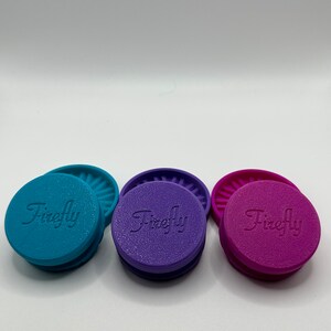Puede incluir: Tres tapas de silicona con la palabra "Firefly" en relieve en la parte superior. Las tapas son de color turquesa, morado y rosa.