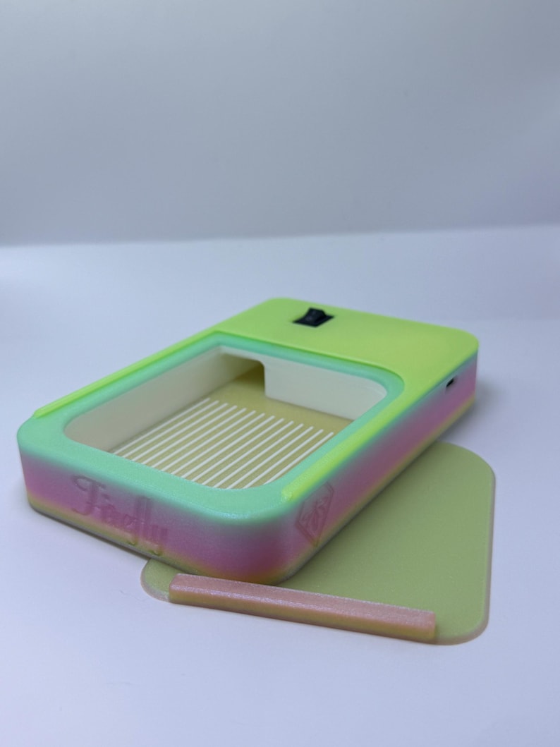 K&ouml;nnte beinhalten: Ein rechteckiges Ger&auml;t in Neongr&uuml;n und Pink mit hellgr&uuml;nem Innenraum und Boden. Das Ger&auml;t hat einen Schalter oben und das Wort "Firefly" ist auf der Seite aufgedruckt. Ein kleiner, hellbrauner Stab liegt auf dem Boden.