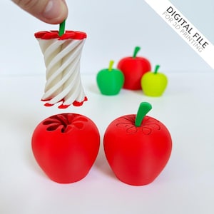 May include: A 3D-printed apple-shaped container with a green stem, next to a white and red apple core. Additional 3D-printed apples in red, green, and yellow are in the background. The image includes the text "DIGITAL FILE FOR 3D PRINTING".