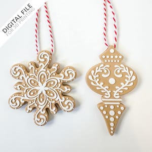 Pode incluir: Dois enfeites de gengibre com desenhos de glacê branco. Um enfeite é um floco de neve e o outro é um enfeite tradicional de árvore de Natal. Ambos os enfeites estão pendurados em um barbante vermelho e branco.