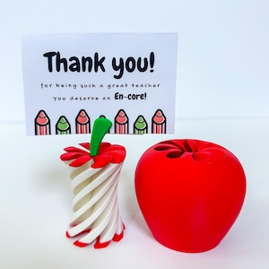 May include: A red apple-shaped pencil holder and a white and red pencil-shaped holder are displayed next to a card that says "Thank you!" in black text. The card also says "for being such a great teacher You deserve an En-core!"