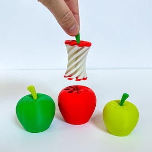 May include: A hand holding a green stem of a white and red apple core above three colorful apple-shaped objects. The apples are green, red, and yellow-green. The core is white with red accents.