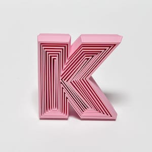 May include: A three-dimensional letter K in a light pink color, featuring a layered, nested design that creates a series of concentric rectangular and angular shapes within the structure of the letter, giving it a textured, stepped appearance.