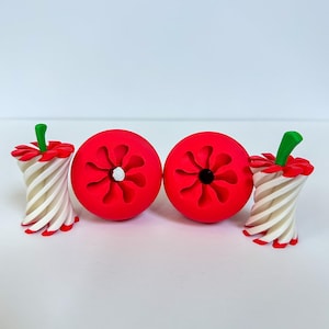 May include: A collection of whimsical, red apple-shaped objects with a flower-like design, paired with candy cane-style stems. The stems are white and red striped, topped with green accents. The objects are arranged on a white surface.