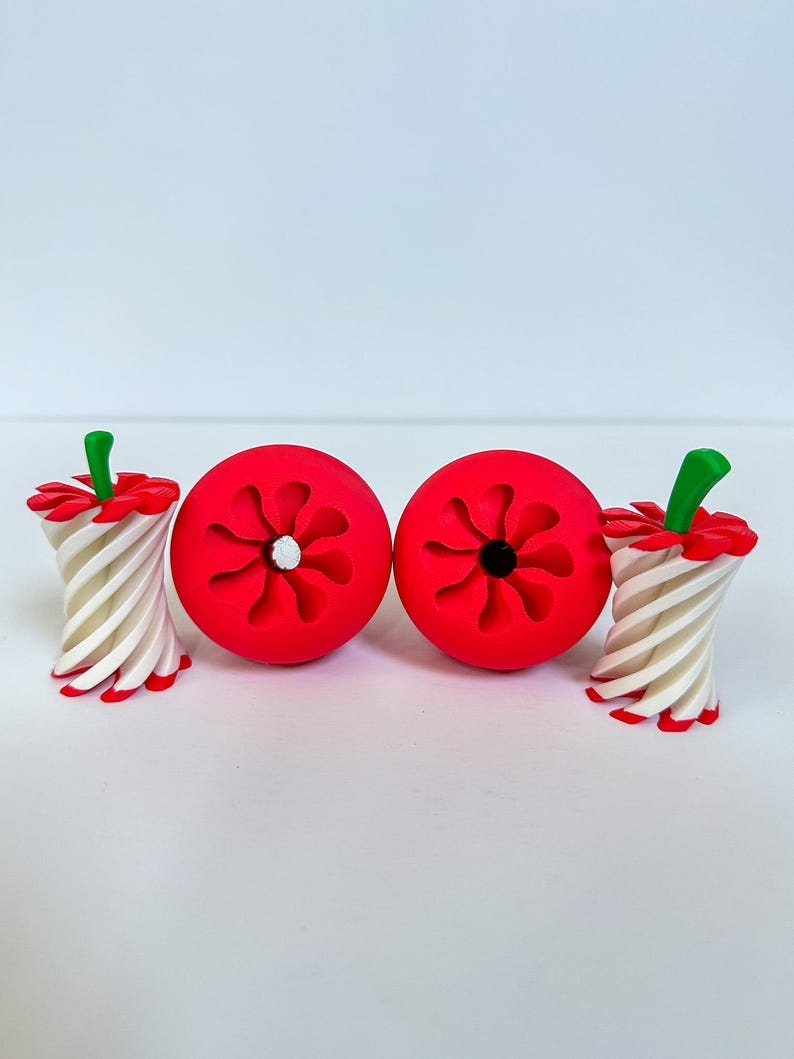 May include: A collection of whimsical, red apple-shaped objects with a flower-like design, paired with candy cane-style stems. The stems are white and red striped, topped with green accents. The objects are arranged on a white surface.
