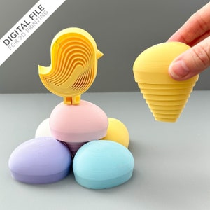 Könnte beinhalten: Ein gelber Vogel mit einem geschichteten Design sitzt auf einem Stapel pastellfarbener Eier. Die Eier sind in den Farben Rosa, Lila, Blau und Gelb gehalten. Der Vogel ist ein 3D-gedrucktes Objekt.