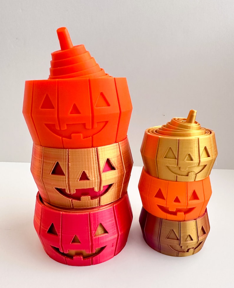 Pu&ograve; includere: Un set di tre contenitori a forma di zucca stampati in 3D, impilati, di colore arancione, oro e rosso. La zucca in alto &egrave; arancione, la zucca al centro &egrave; dorata e la zucca in basso &egrave; rossa. Ogni zucca ha un viso di zucca intagliato.
