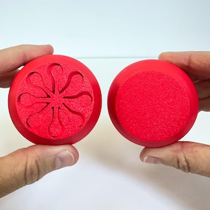 May include: Two red, circular objects are held up against a white background. One has a textured surface, while the other features a carved floral design. The objects appear to be bottle stoppers or decorative elements.