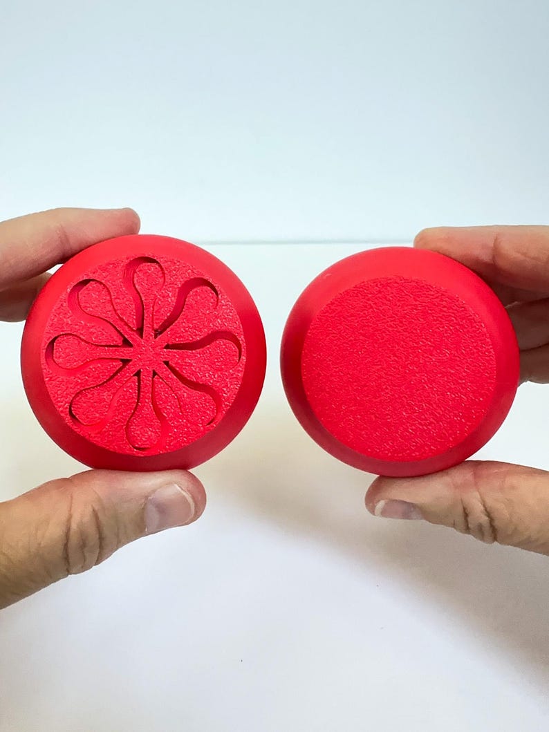 May include: Two red, circular objects are held up against a white background. One has a textured surface, while the other features a carved floral design. The objects appear to be bottle stoppers or decorative elements.