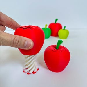 May include: A red apple-shaped kitchen tool with a white and red spiral base, held by a hand. Several other apple-shaped objects in red, green, and yellow are in the background. The tool is designed for coring apples.