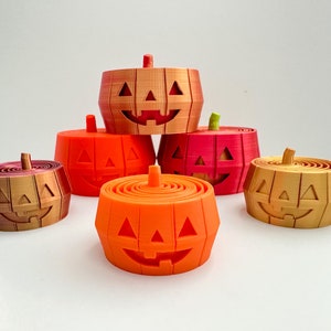 Pu&ograve; includere: Cinque contenitori a forma di zucca di Halloween di colore arancione, rosso e oro con un viso sorridente intagliato. I contenitori sono impilati uno sopra l'altro.
