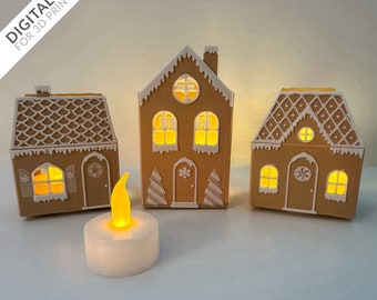 Può includere: Tre portacandele di carta a forma di case di pan di zenzero con glassa bianca e finestre. Le case sono illuminate dall'interno con candele tealight. Le case sono su una superficie bianca.