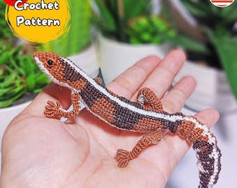 May include: Crocheted lizard amigurumi toy with brown, white, and black stripes. The lizard is posed on an open hand, with its tail curled around the hand.