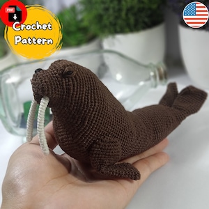 May include: A brown crocheted walrus amigurumi toy with white yarn for the tusks. The toy is being held in a person's hand. Crochet Pattern PDF.